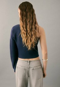 Marineblauer, kurz geschnittener Pullover mit einem langen Ärmel, kombiniert mit hellgrauen, hochtaillierten Jeans. Das Model hat lange, wellige Haare. Minimalistischer Stil.