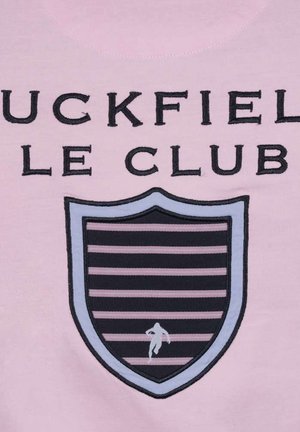 Pink stof med broderet tekst "UCKFIELD LE CLUB" over et skjoldformet emblem med vandrette sorte og lyserøde striber samt en lille løbende figur.