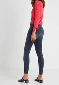 Vaqueros ajustados de mezclilla azul oscuro, con una silueta ceñida, combinados con una blusa de manga larga en color coral brillante que presenta un detalle de lazo en los puños. Zuecos negros con tachuelas.