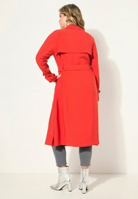 Rode trenchcoat met een riem aan de achterkant en zijsplitten, gemaakt van een soepel materiaal. Gepaard met zilveren enkellaarzen, gedragen bovenop grijze strakke broek.