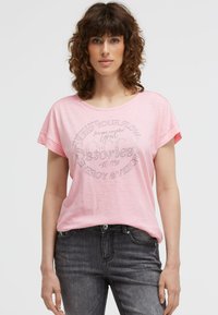 Woman with curly brown hair wearing a pink T-shirt with text and dark gray jeans, standing against a plain light background.