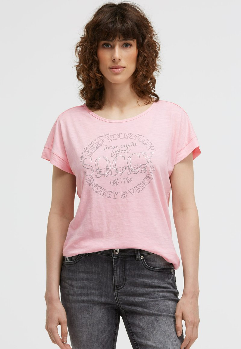 Woman with curly brown hair wearing a pink T-shirt with text and dark gray jeans, standing against a plain light background.
