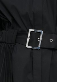 Melnas auduma apģērbs ar plisu dizainu, kas aprīkots ar platu jostu. Jostai ir spīdīgs sudraba kvadrātveida aizdare ar iegravētu uzrakstu "KARL LAGERFELD".