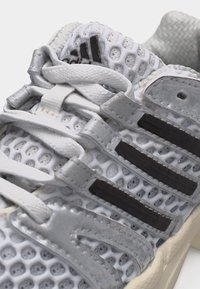 Close-up of white and gray Adidas sneaker with mesh upper, black stripes, and white laces tied on top.