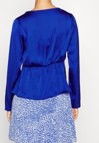 Blusa in satin blu con maniche lunghe, vita elastica e design a peplo. Tessuto liscio, scollo rotondo e silhouette fluida.
