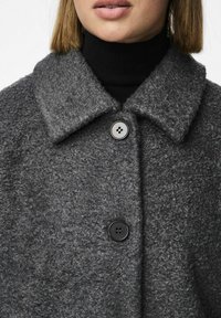 Cappotto grigio texturizzato con un ampio colletto e tre bottoni neri. Indossato su un dolcevita nero a coste. Materiale morbido e accogliente.