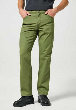 Pantalones de algodón verdes con corte recto, que cuentan con bolsillos delanteros y una cremallera en la parte frontal. Se llevan con calzado negro, creando un look casual.