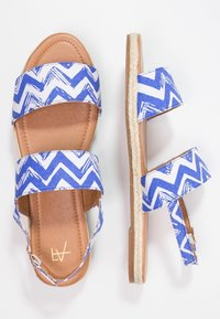 Blå og hvide mønstrede sandaler med to brede stofstropper, en tan faux læder fodseng og en jute reb sål. Justerbar ankelrem inkluderet.