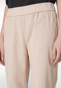 Housut vaaleanbeigellä värillä, kevyet ja joustavalla vyötäröllä, joissa on sileä pinta ja rento istuvuus, osittain näkyvissä kuvassa.