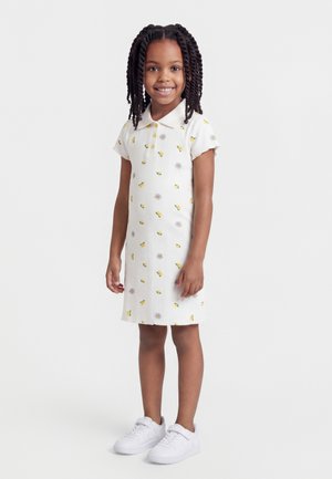 Mladé dievča sa usmieva, stojí, oblečené v bielych polokošeľových šatách so žltými kvetinovými vzormi a bielych teniskách na jednoduchom pozadí.