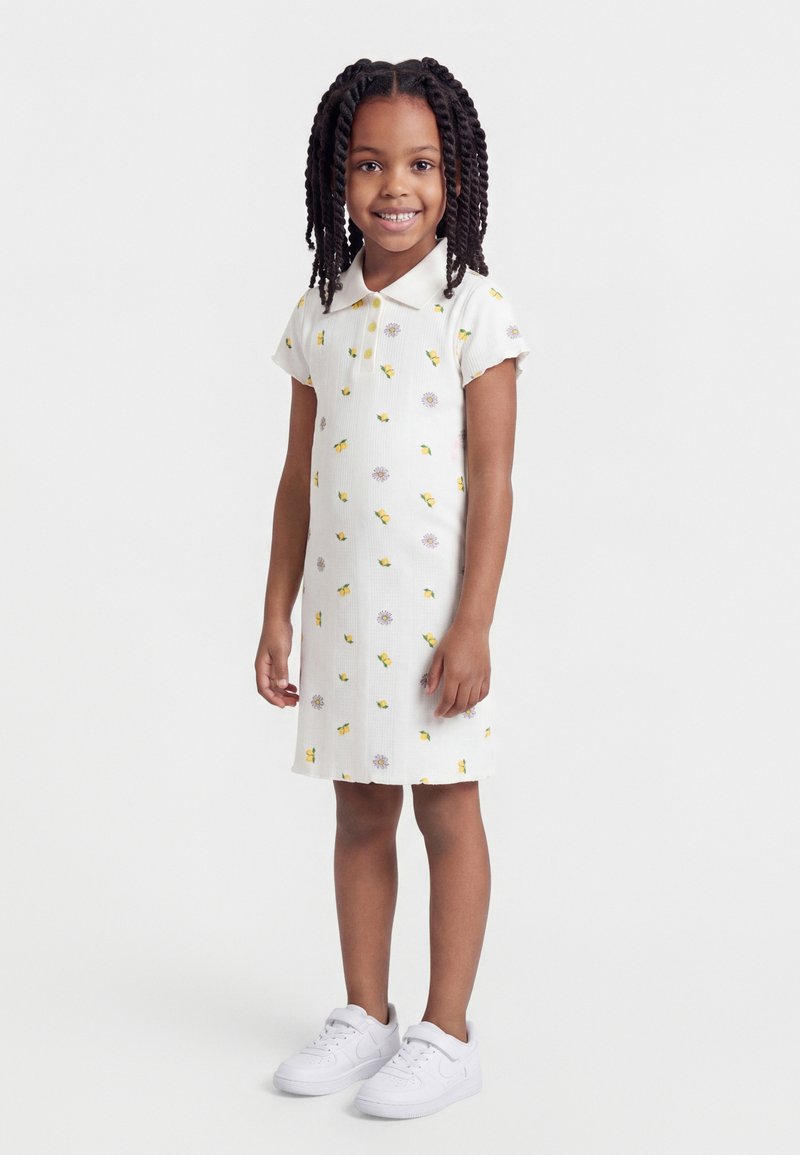 Νεαρή κοπέλα που χαμογελά, στέκεται, φορώντας ένα λευκό φόρεμα πόλο με κίτρινα λουλούδια και λευκά αθλητικά παπούτσια σε απλό φόντο.