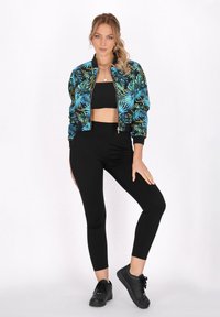 Zwarte bomberjack met blauw en groen tropisch bladpatroon, gecombineerd met een zwarte crop top en hoge zwarte legging. Zwarte sneakers.