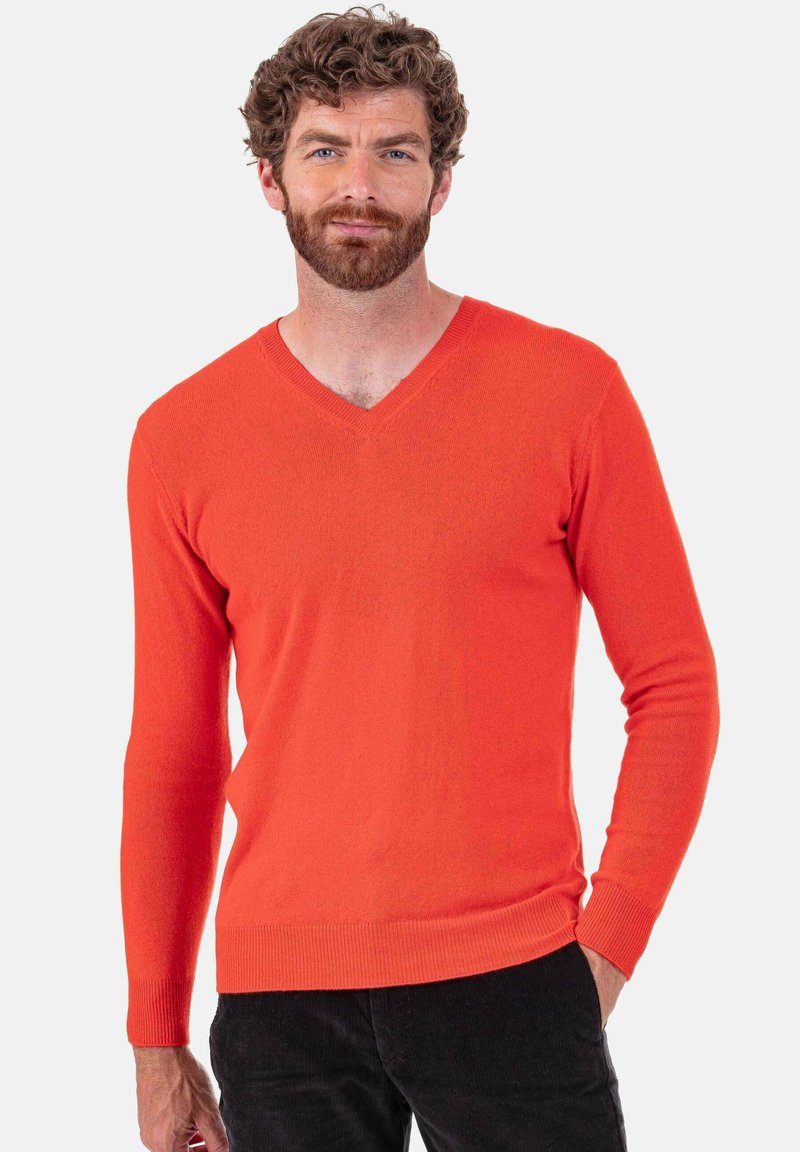 Pull orange à col en V avec manches longues, poignets et ourlet côtelés. Tissu doux et texturé avec une coupe ajustée. Pantalon noir visible en dessous.