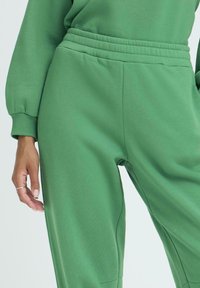 Zelene pamučne trenirke s elastičnim pojasom, bočnim džepovima i opuštenim krojem. Glatka tekstura i minimalistički dizajn.