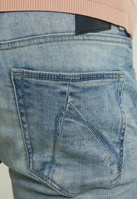 Blå denimjeans med en blekt textur, med en unik design på bakfickan och en svart läderlapp vid midjan.