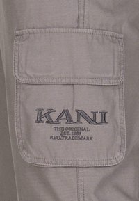 Tissu gris avec une poche appliquée. Logo "KANI" brodé en noir, accompagné de "THE ORIGINAL EST. 1989 MARQUE DÉPOSÉE" en texte plus petit en dessous.