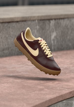 Donkerbruine leren sneaker met beige Nike-vink, beige veters en gumzool zwevend boven stenen oppervlak in een buitenomgeving.