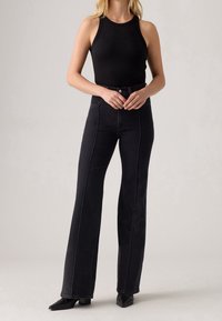 Femme portant un haut noir côtelé sans manches, rentré dans un jean noir taille haute à jambes larges, avec des chaussures noires pointues.