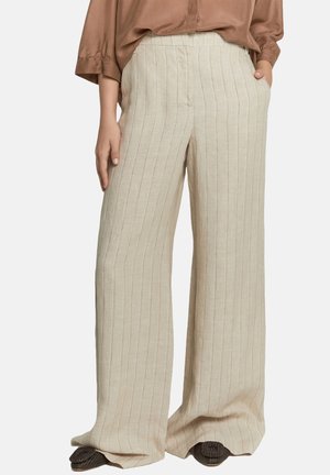 Pantaloni beige a gamba larga con sottili righe verticali, indossati con una camicetta marrone e scarpe intrecciate scure. Mani nelle tasche e lungo i fianchi.