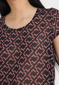 Top noir transparent avec un motif géométrique rouge et blanc complexe, doté d'un col rond et de manches courtes.