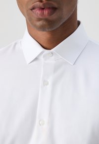 Zbliżenie na dolną część twarzy i szyi osoby noszącej białą koszulę zapinaną na guziki z klasycznym kołnierzem.