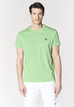Uomo che indossa una maglietta verde chiaro a maniche corte e pantaloncini bianchi, in piedi davanti a uno sfondo chiaro e uniforme.