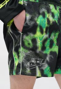 Main d'une personne dans la poche d'un short noir avec un motif camouflage numérique vert et gris et le logo blanc Nike Air sur la jambe.
