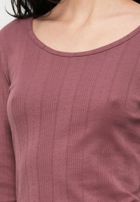 Μώβ μπλούζα με μακριά μανίκια και στρογγυλή λαιμόκοψη. Διαθέτει ριμπ υφή με κάθετες ρίγες και μικρά διακοσμητικά σχέδια σε όλη την επιφάνεια.