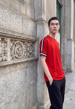 Jeune homme en chemise rouge avec des rayures noires et blanches sur les manches, appuyé contre un mur en pierre orné, regardant sur le côté.