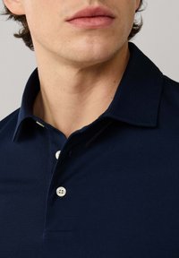 Primer plano de la parte inferior del rostro y cuello de un hombre que lleva una camisa tipo polo azul marino con botones blancos y cuello vuelto hacia abajo.