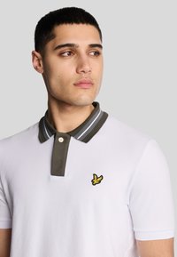 Hvid polo t-shirt med en mørkegrøn krave, der har grå striber. Indeholder et lille gult broderet logo på venstre bryst.