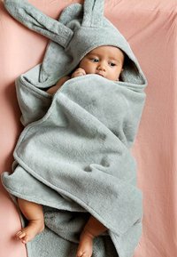 Grå, huva handduk med kaninöron, tillverkad av mjuk, plyschig tyg. Handduken sveper runt en delvis synlig spädbarn, som vilar på en rosa yta.