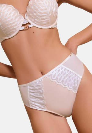 Naine, kes kannab beeži pesukomplekti läbipaistvate mustriliste pitsdetailidega rinnahoidjal ja pükstel, poseerib ühe käega puusal.