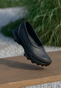 Sorte slip-on sko laget av glatt skinn med teksturerte såler, rund design og en stroppe bak på hælen for enkel påkledning.