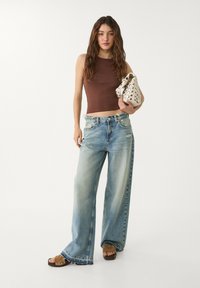 Ljusblå, vida jeans i denim, kombinerade med en brun åtsittande croptop. Modellen håller en vit väska med cirkulära utskärningar och nitar.