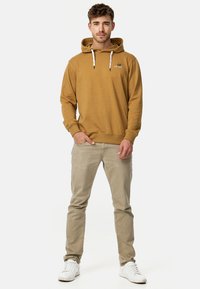 Brązowa bluza z kapturem i białymi sznurkami, z detalem logo na klatce piersiowej, zestawiona z jasnobeżowymi spodniami i białymi sneakersami. Prosty, casualowy strój.