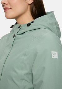 Femme portant une veste à capuche légère verte avec des boutons-pression noirs et un petit écusson blanc sur le bras supérieur.