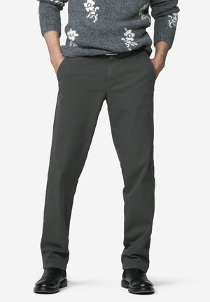 Mann trägt dunkelgraue gerade geschnittene Hose, schwarze Anzugschuhe und einen grauen Pullover mit weißen Blumenmustern, Hände in den Taschen.