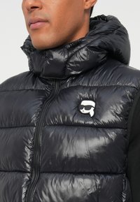 Melna puffa veste ar augstu apkakli, ar spīdīgu tekstūru, horizontālu izšūtu dizainu un baltu logotipa plāksni kreisajā krūtī.