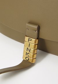 Oljčno zelena usnjena torba s gladko teksturo, z zlatim kovinskim okrasom in vtisnjenimi črkami, ki črkujejo "PINKO."
