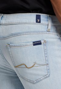 Jasnoniebieskie dżinsy mają małą metkę marki na tylnej kieszeni oraz złotą, falistą stępnę.