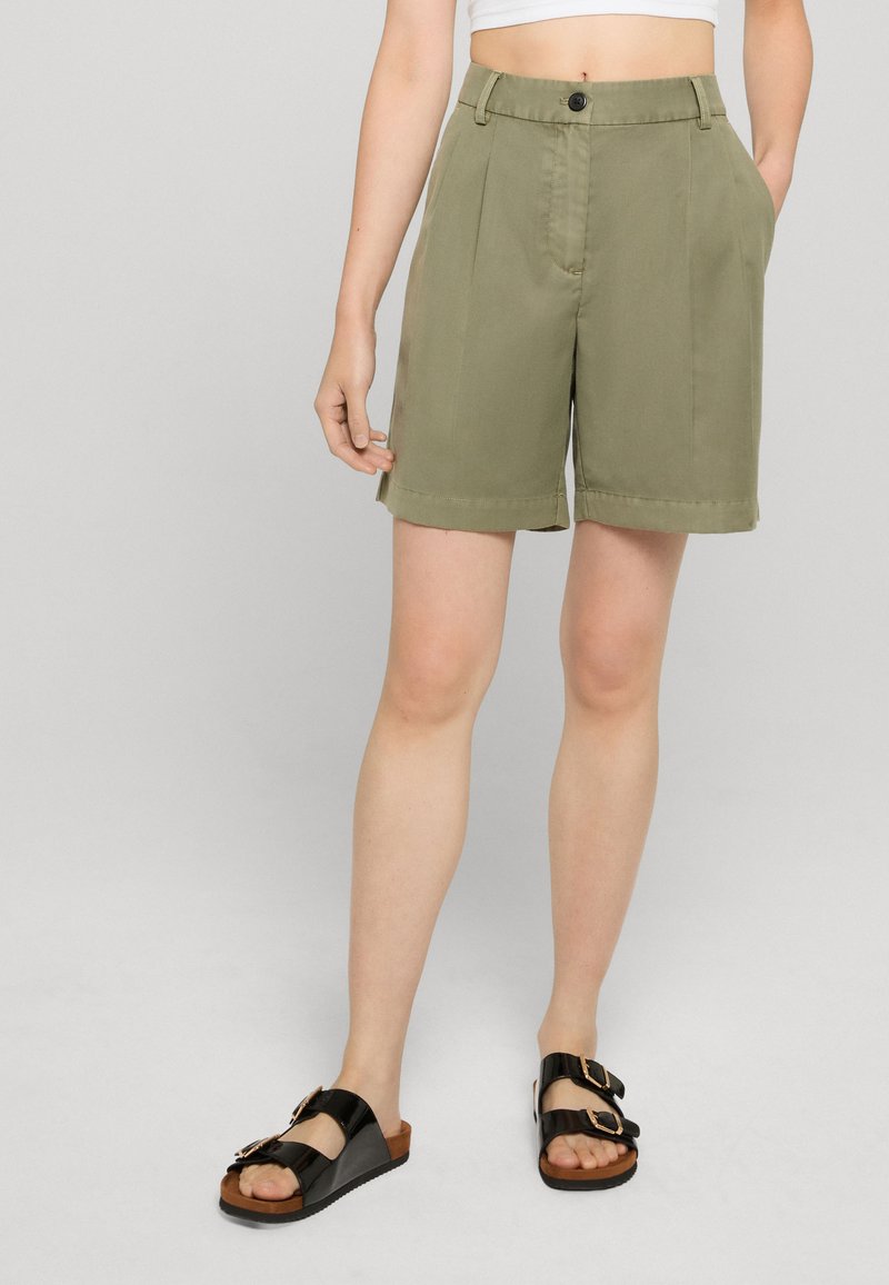 Personne portant un short taille haute vert olive avec des plis sur le devant et des sandales noires à double brides, debout devant un fond clair.