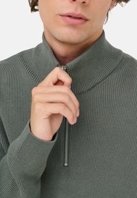 Grønneribbet sweater med høj krave og halv lynlås. Stoffet ser tekstureret ud, og lynlåsen har en metallisk finish. Tæt billede af en hånd, der holder lynlåsen.
