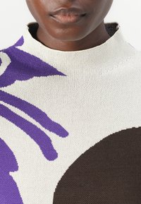 Knitted sweater featuring a high neckline, with a cream base, purple botanical patterns, and a dark brown circular accent. Soft texture.