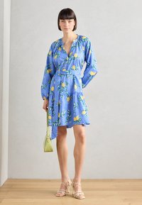 Rochie albastră cu imprimeu de lămâie, mâneci lungi, decolteu în V și curea de legat. Este purtată cu sandale bej cu curea și o geantă mică verde. Fundal din podea din lemn.