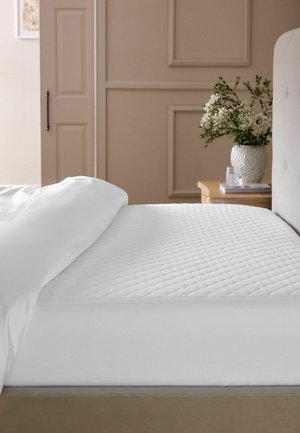 Materasso bianco con copertura trapuntata su struttura del letto beige, parzialmente coperto da piumone bianco, accanto a un comodino in legno con vaso di fiori bianchi.