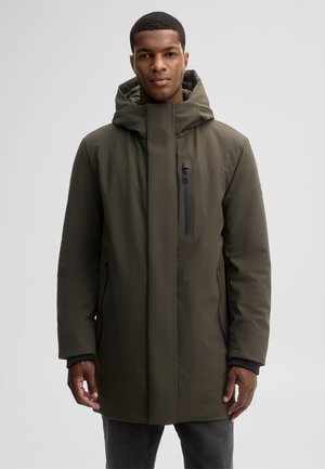 Man wearing an olive green hooded winter coat with zipped pockets, standing against a plain light gray background.