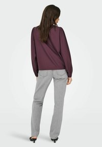 Vrouw in een paars blouse met lange mouwen, lichte grijze jeans en zwarte hakken, staand met haar rug naar de camera op een witte achtergrond.