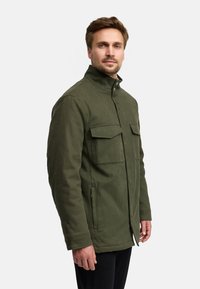 Olijfgroene jas met een hoge kraag, twee borstzakken en zijritsen. De stof heeft een gestructureerde afwerking, ontworpen voor warmte en praktische gebruik.