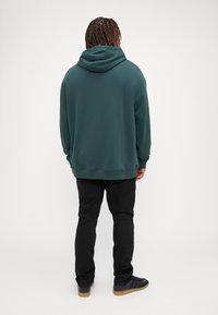 Sudadera verde oscura con un bolsillo trasero, puños y dobladillo acanalados. Combinada con pantalones negros y zapatos negros con una suela texturizada.